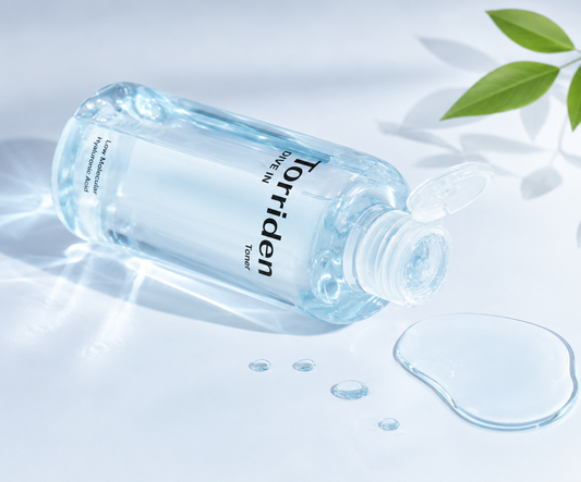A clear plastic bottle of Torriden Water Toner lying on its side with the cap open, showing transparent light blue liquid inside. A small puddle of the clear toner is placed beside it on a bright white surface with soft leaf shadows and a green leaf in the corner.